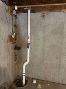 A sump pump system in a basement with exposed concrete walls. PVC piping runs vertically from the sump pit in the floor, connecting to a wall-mounted pipe and electrical components nearby.
