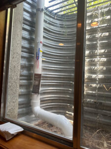View through a basement window showing a curved white pipe running vertically and then bending at a right angle, inside a corrugated metal window well with a metal grate and some leaves on the ground.