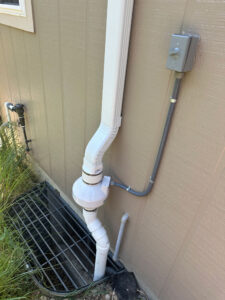 A white PVC radon mitigation system is installed on the exterior wall of a house, with pipes running vertically and a fan unit, next to a metal window well grate and an electrical control box.
