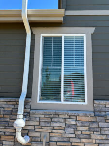A rectangular window with white blinds is set in a dark green wall with tan trim. There is a white downspout attached to the wall and a stone facade at the base of the house.