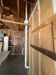 A garage with unfinished walls, exposed wooden beams, and shelves. A white PVC pipe runs vertically along the wall, and an orange extension cord hangs nearby. A brown door is at the back.