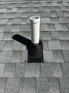 A white vent pipe with a protective cap extends from a gray shingled roof. The pipe is surrounded by a black flashing base, and it casts a shadow to the left on the roof.